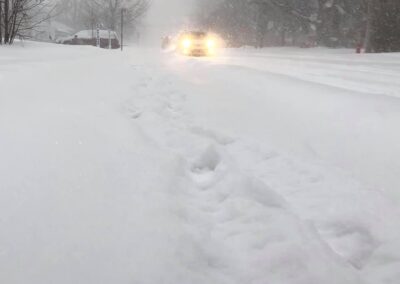 Car with headlights driving down a snow-covered road near sidewalk with falling snow